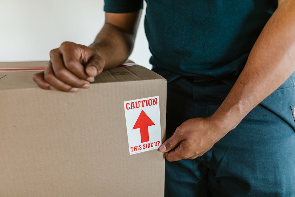 Two women engaged in a home relocation process are standing indoors surrounded by stacked cardboard boxes, some sealed with red and black striped packing tape. One woman is holding a medium-sized box