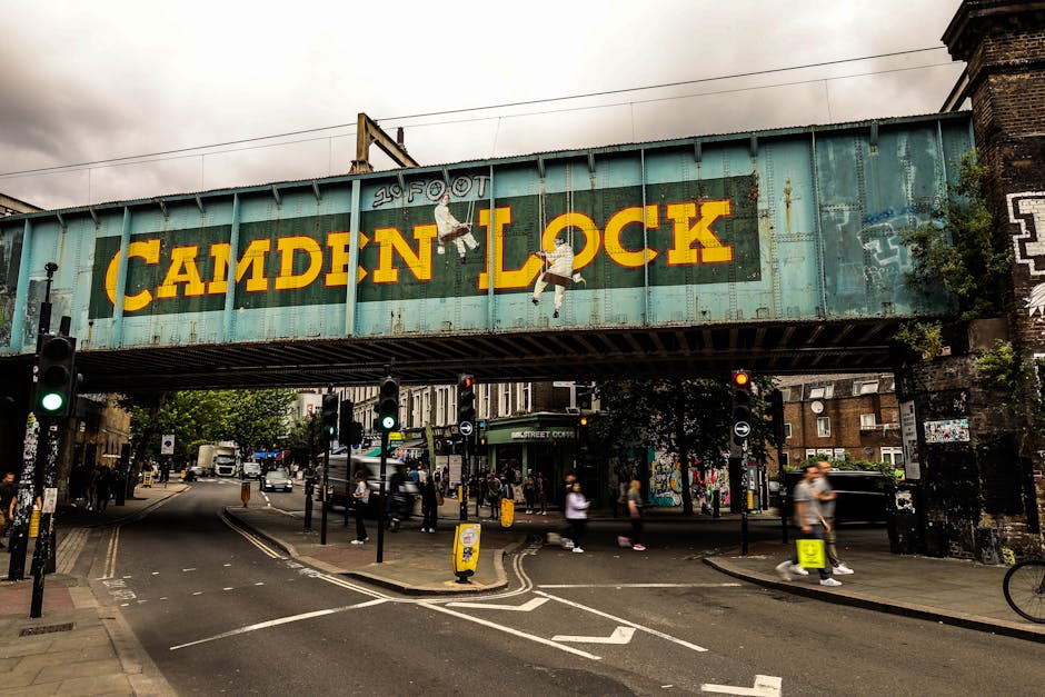 A professional moving team from Self Storage Clapham is indoors in a well-lit room with large, arched windows. One mover, standing and smiling, is carrying two cardboard boxes stacked one on top of th